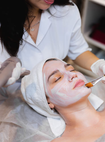 beautician applying white moisturizer mask on the face of a girl client in spa beauty salon.